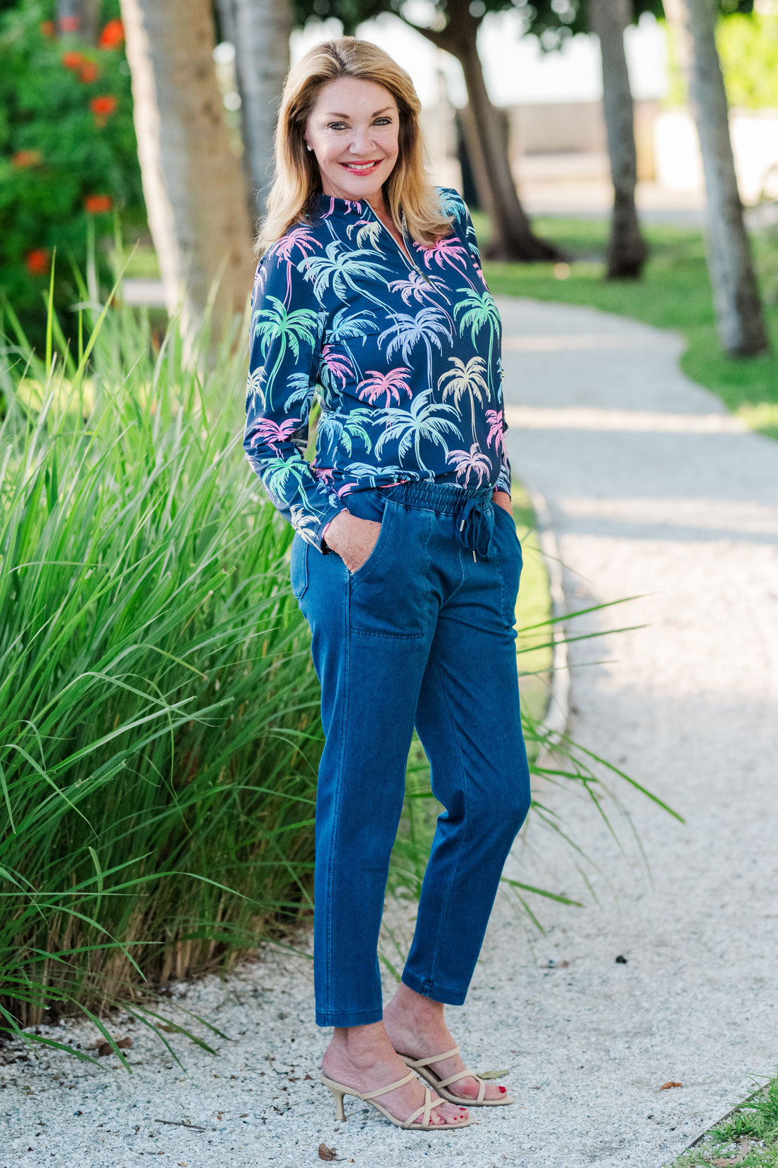 Woman wearing a colorful palm tree patterned shirt and blue pants standing outdoors.