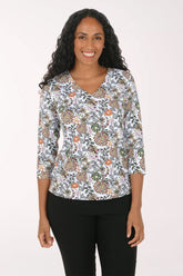 A woman with long curly black hair wearing a white three-quarter sleeve top with a colorful floral and paisley pattern, paired with black pants, standing against a plain white background.