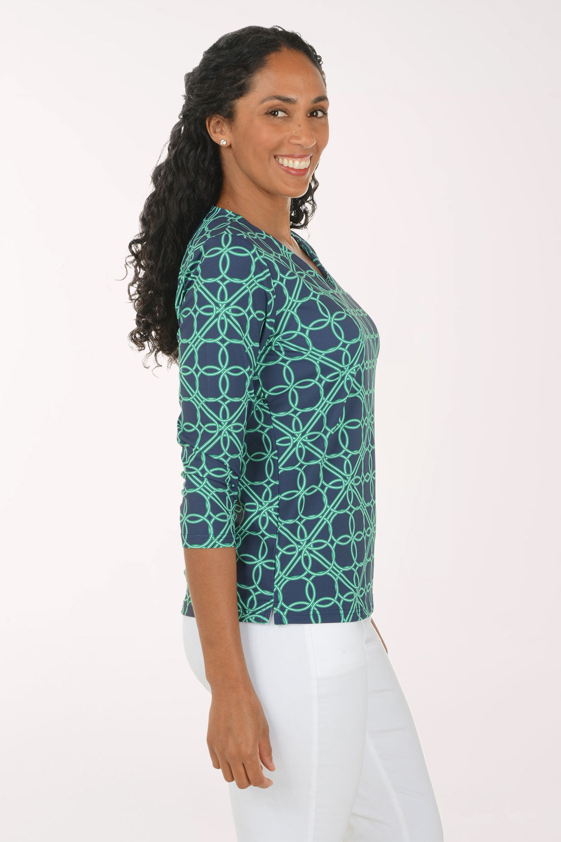 A woman modeling a three-quarter sleeve V-neck top with a teal and navy geometric interlocking circle pattern, paired with white pants, against a plain white studio background.