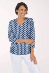 A woman modeling a blue and white geometric print long-sleeve top with a V-neck, paired with white pants, against a plain light background.