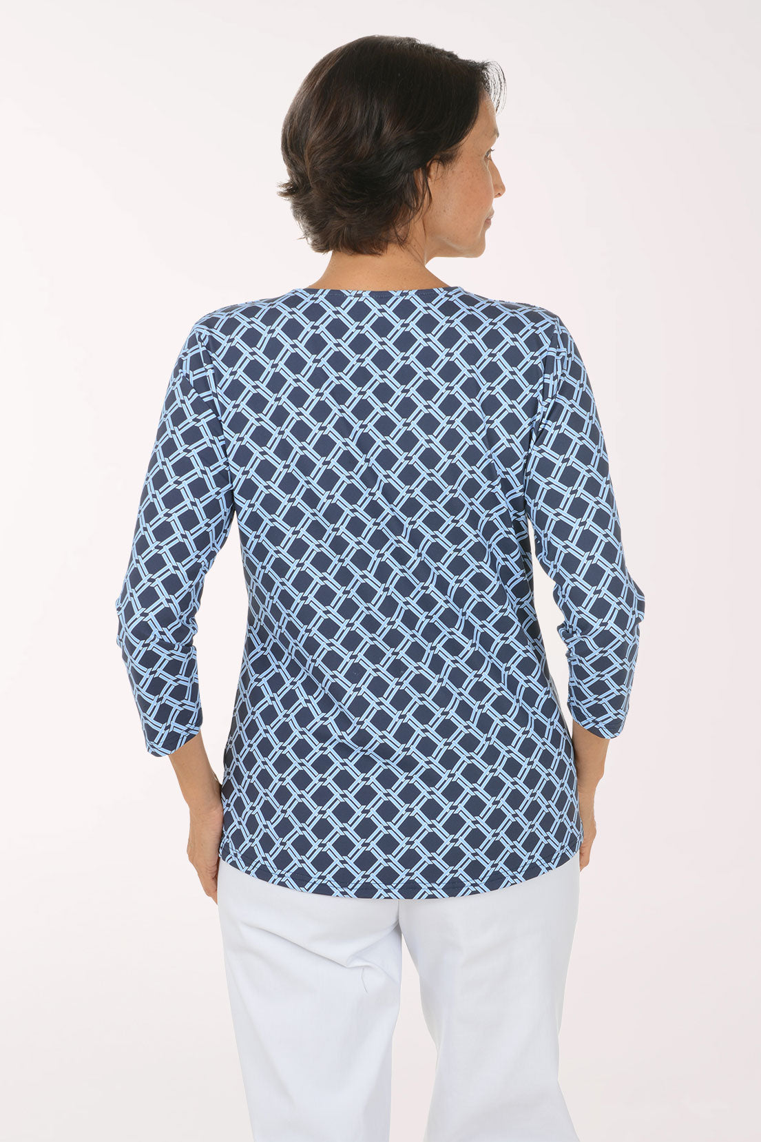 A woman modeling a blue and white geometric print long-sleeve top with a V-neck, paired with white pants, against a plain light background.