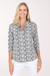 Front view of a model wearing the Anthony's Resort Wear Neutral Chevron UPF50+ Ruffle Neck Top in a black and white brushstroke chevron print, with a ruffle-trimmed V-neckline and 3/4 sleeves, worn with white pants.