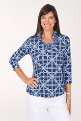 Front view of a model wearing the Anthony's Resort Wear Blue Lattice Rings UPF50+ Ruffle Neck Top, a navy top with a light blue interlocking ring lattice print, ruffle-trimmed V-neckline and 3/4 sleeves, worn with white pants.