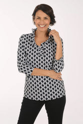 A woman modeling a black and white geometric patterned blouse with three-quarter sleeves and a V-neck, paired with black pants, against a plain white studio background.