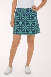 A woman modeling a short A-line skirt with a teal and navy geometric interlocking circle pattern, paired with a white top and white slip-on shoes, against a light studio background.