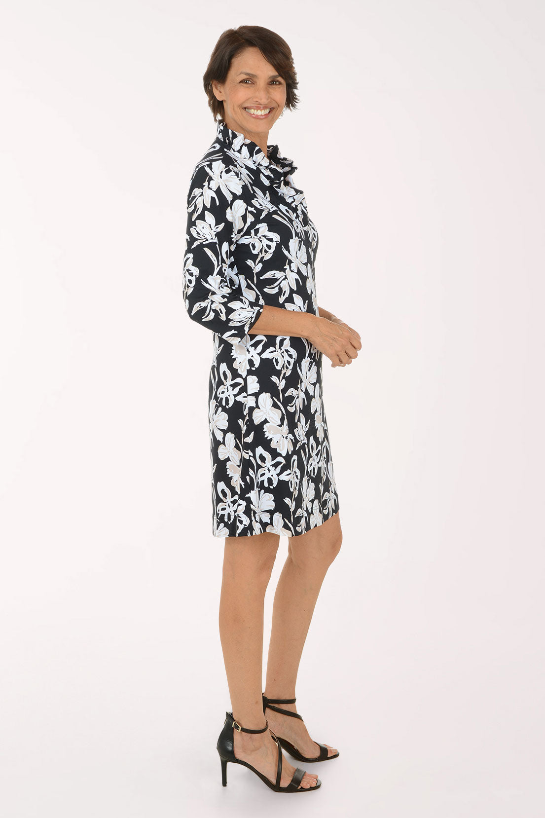 A woman modeling a black and white floral print shirt dress with three-quarter sleeves and a collared neckline, paired with black strappy high-heeled sandals against a plain white studio background.