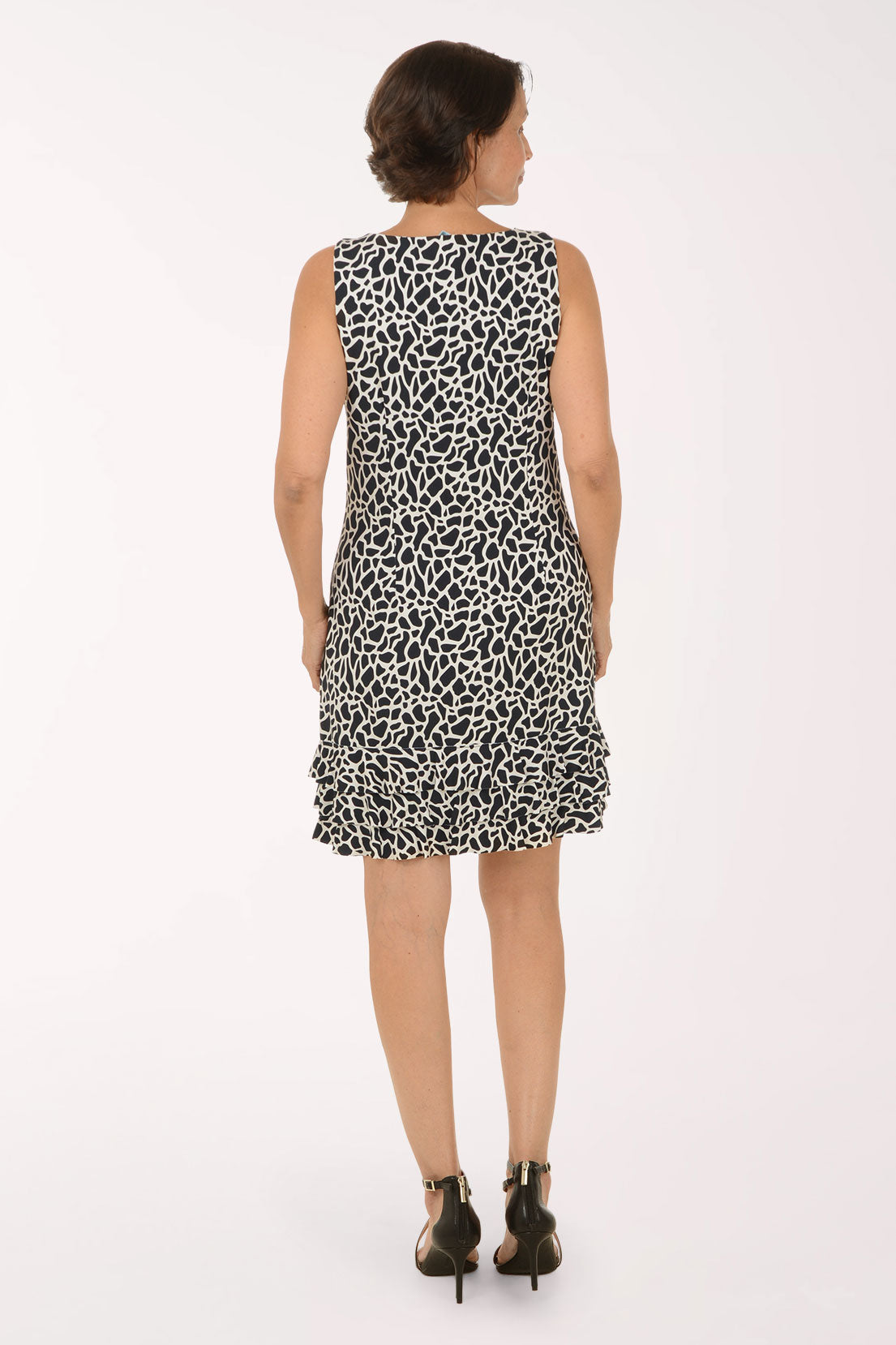 A woman modeling a sleeveless black and white animal print dress with a ruffled hem, paired with black strappy high-heeled sandals, against a plain white studio background.