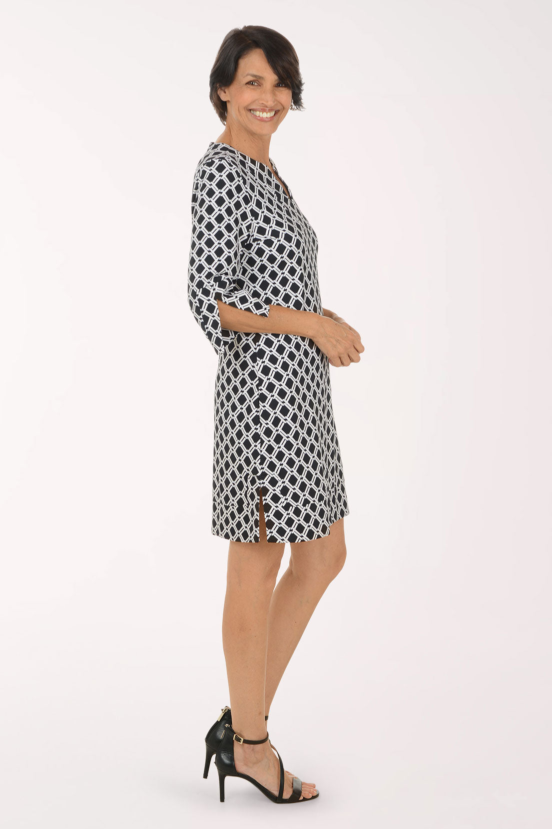 A woman modeling a short black and white geometric patterned dress with elbow-length sleeves and a split neckline, paired with black strappy high-heeled sandals, against a plain white studio background.