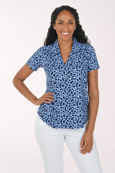 A woman modeling a short-sleeve collared blouse with a blue and white abstract animal-like print, paired with white pants, against a plain light background.