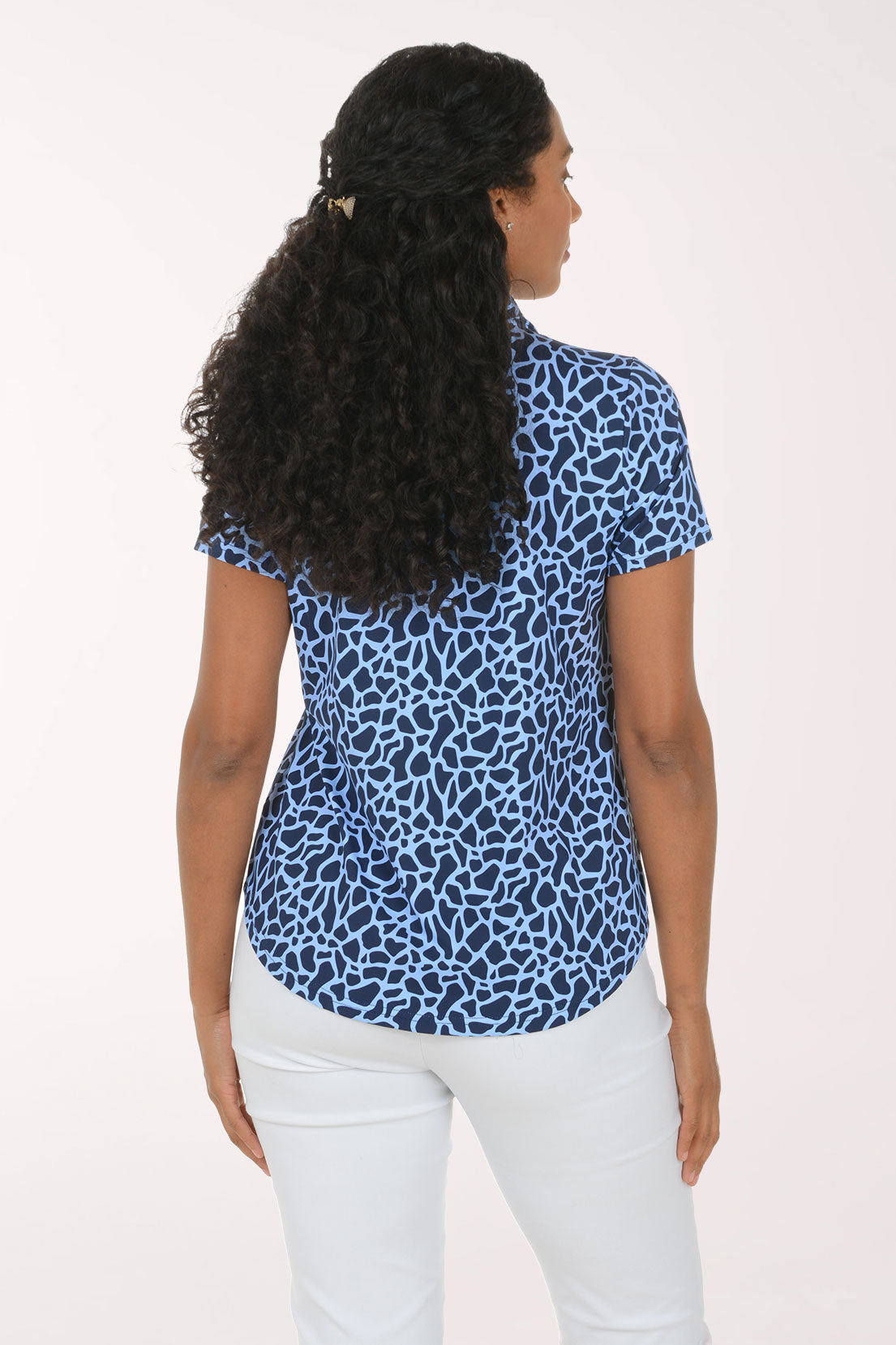 A woman modeling a short-sleeve collared blouse with a blue and white abstract animal-like print, paired with white pants, against a plain light background.