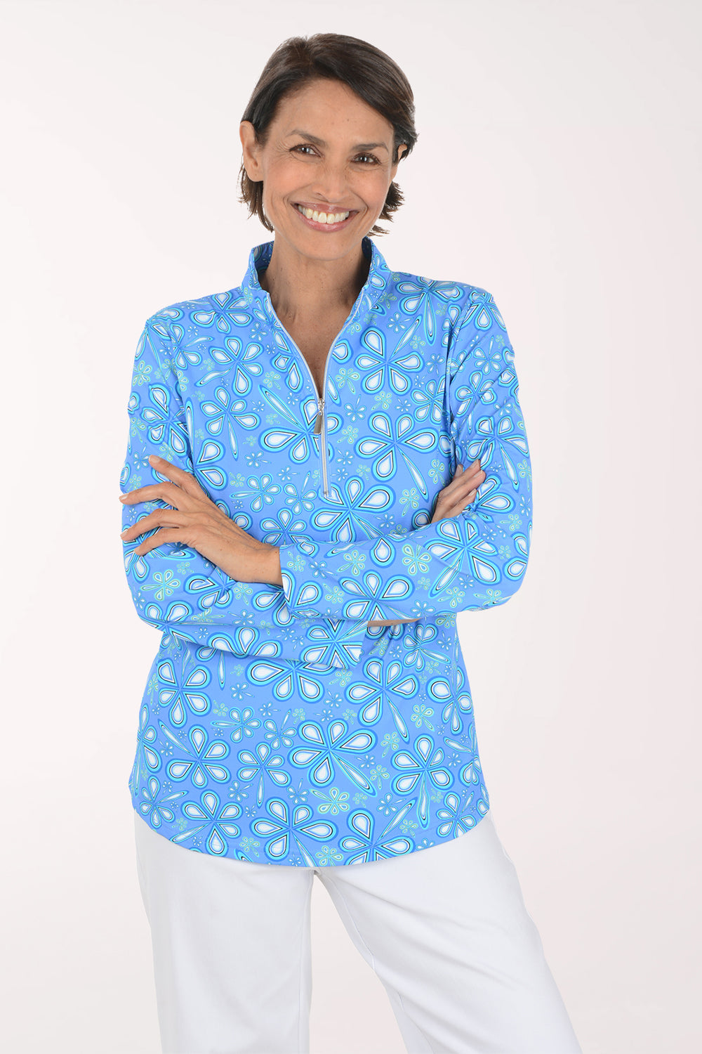 A smiling woman with short brown hair stands against a light off-white studio background, wearing a light blue long-sleeve quarter-zip pullover with a white paisley pattern and white pants.