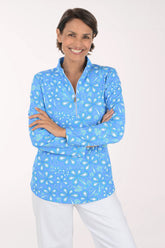 A smiling woman with short brown hair stands against a light off-white studio background, wearing a light blue long-sleeve quarter-zip pullover with a white paisley pattern and white pants.