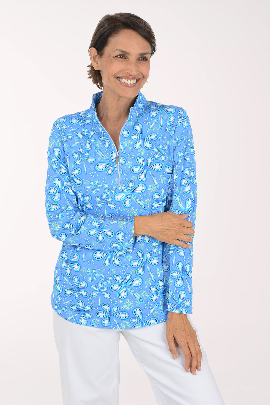 A smiling woman with short brown hair stands against a light off-white studio background, wearing a light blue long-sleeve quarter-zip pullover with a white paisley pattern and white pants.