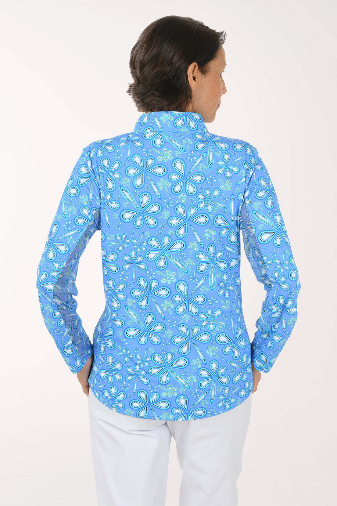 A smiling woman with short brown hair stands against a light off-white studio background, wearing a light blue long-sleeve quarter-zip pullover with a white paisley pattern and white pants.