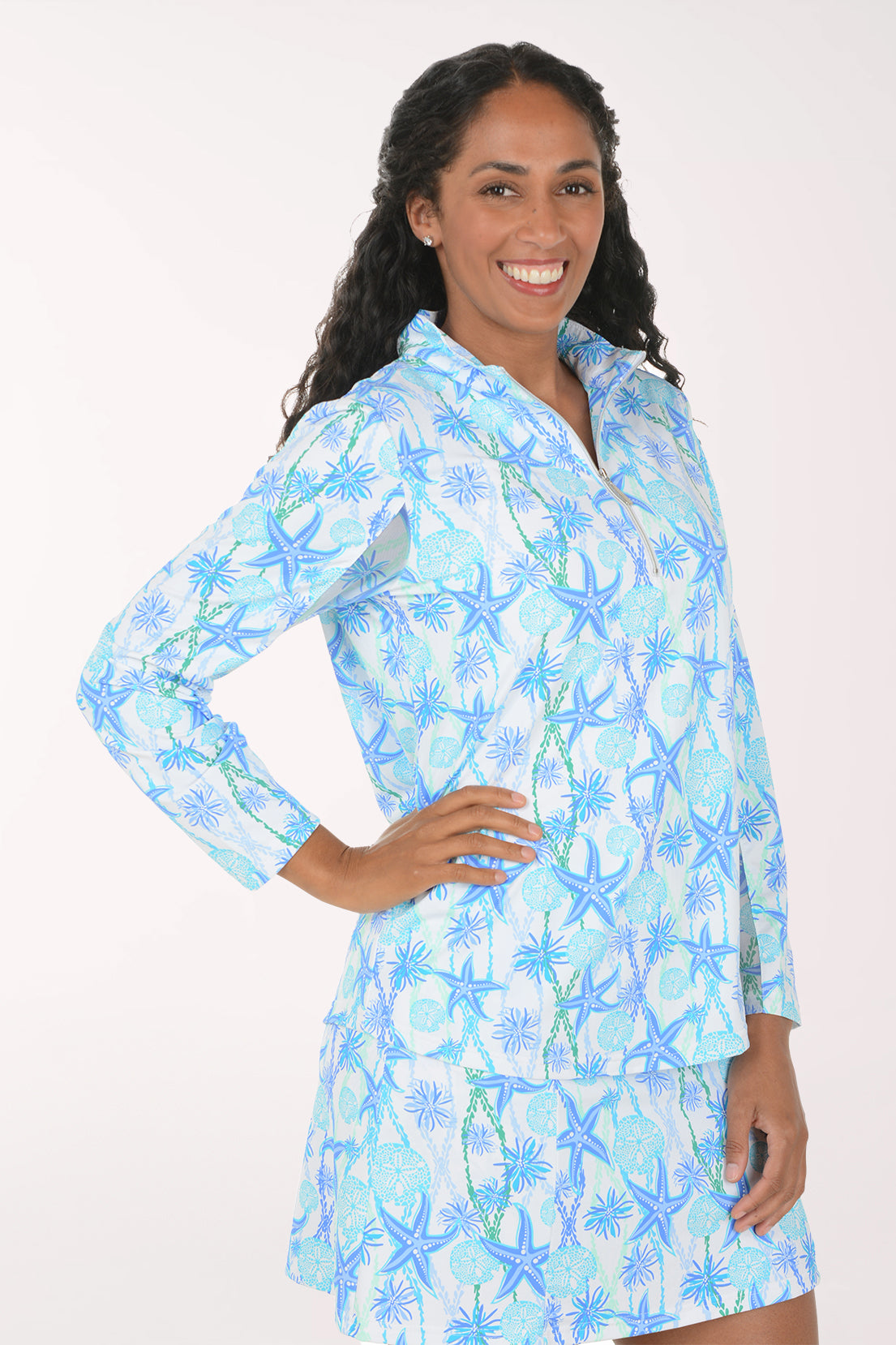 A smiling woman wearing a matching two-piece lounge set with a light blue and white starfish and palm tree print, featuring a long-sleeve quarter-zip top and a matching skirt.