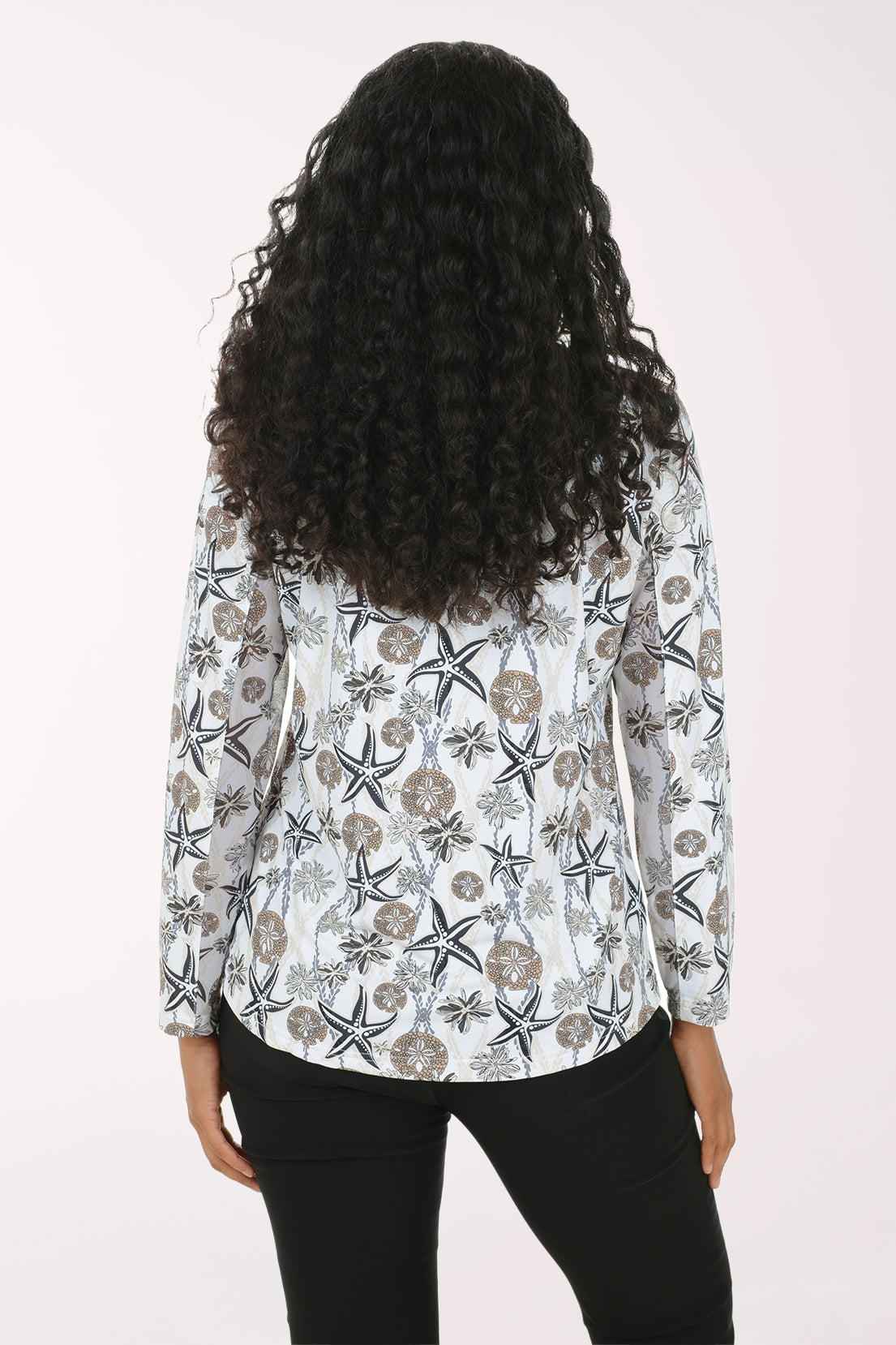 A woman with long, curly black hair is shown from behind wearing a white long-sleeve blouse with a brown and gray starfish pattern, paired with black pants, against a plain light background.
