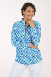A woman modeling a blue and light blue diamond-patterned quarter-zip pullover top with white pants, set against a plain white studio background.