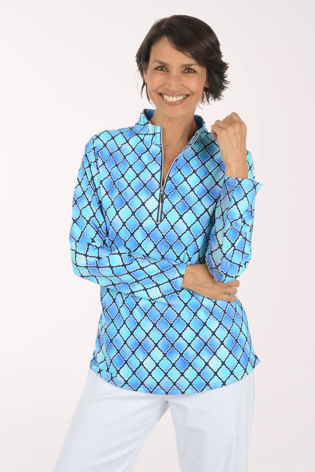 A woman modeling a blue and light blue diamond-patterned quarter-zip pullover top with white pants, set against a plain white studio background.