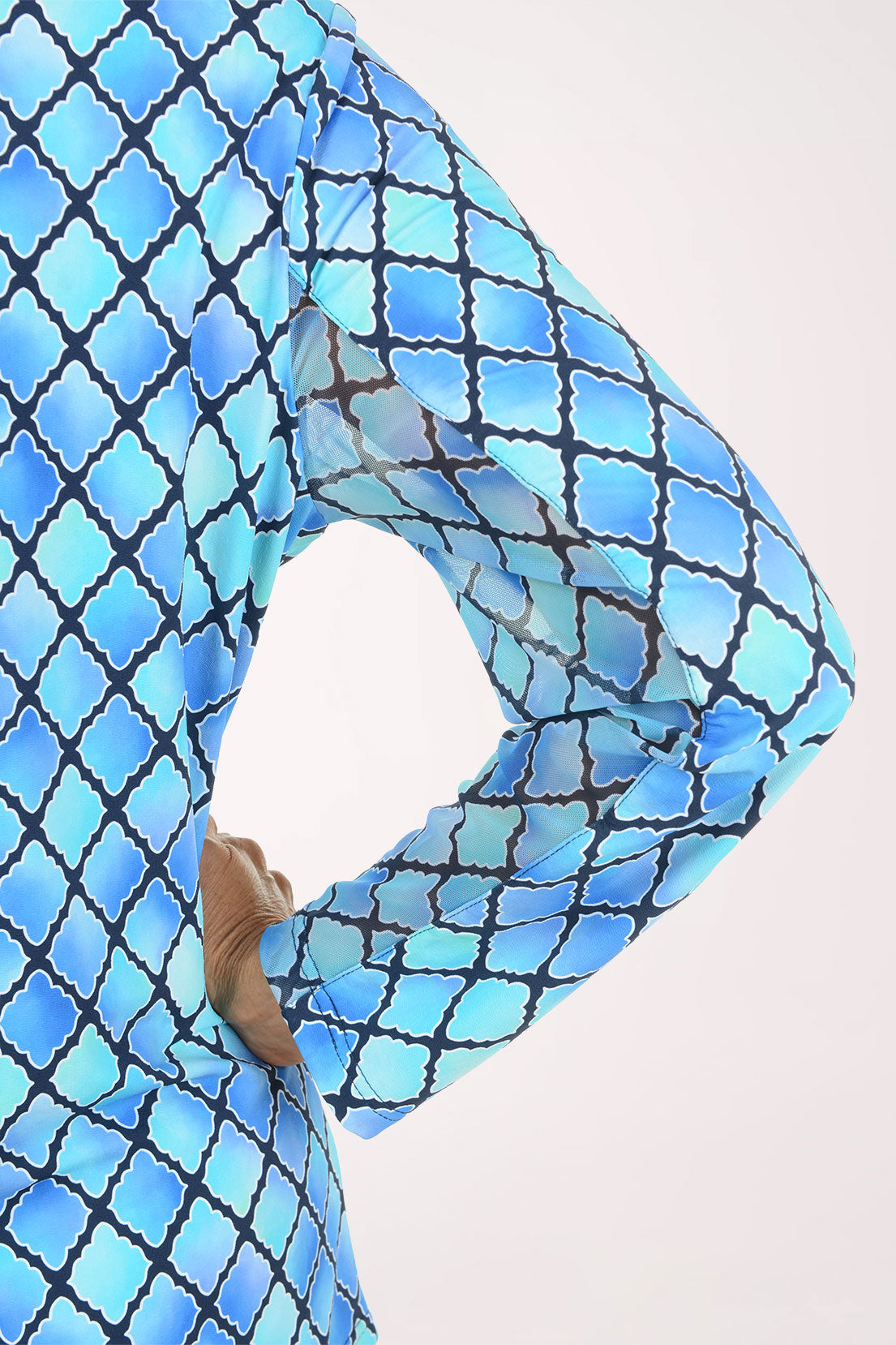 A close-up of a person wearing a sheer long-sleeve top with a blue and light blue Moroccan trellis pattern outlined in black, with a hand tucked into the side pocket.