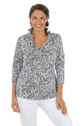 Woman wearing a black and white patterned blouse with white pants on a white background.