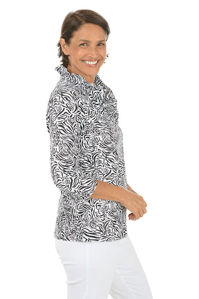 Woman wearing a black and white patterned blouse with white pants on a white background. Side shot.