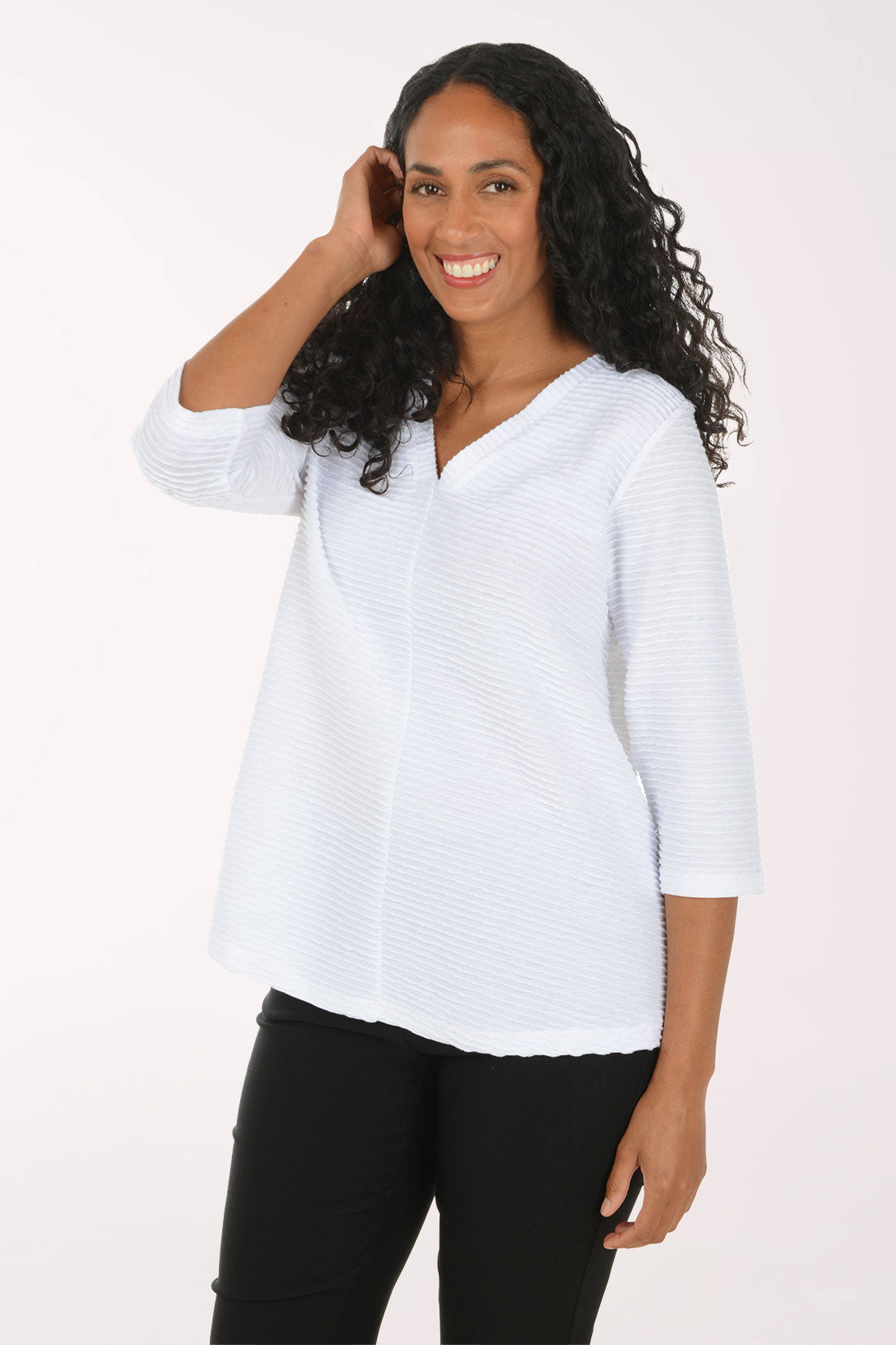 A woman with long curly black hair wearing a white textured three-quarter sleeve top and black pants, posing against a plain white studio background.
