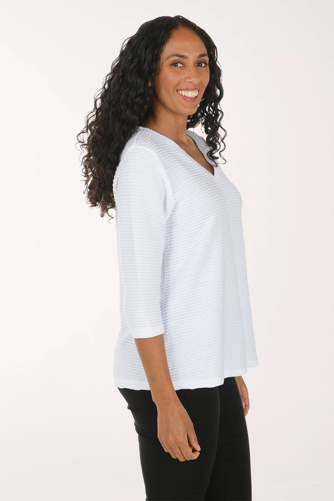 A woman with long curly black hair wearing a white textured three-quarter sleeve top and black pants, posing against a plain white studio background.