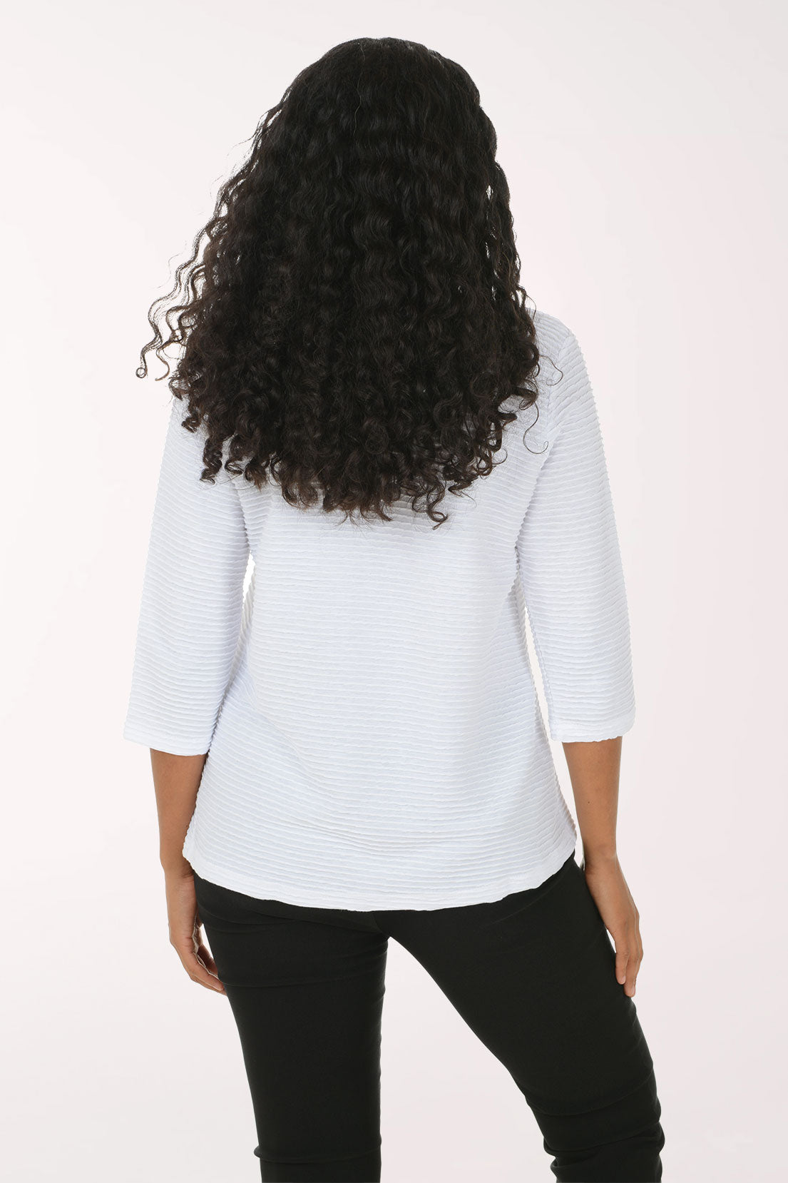 A woman with long curly black hair wearing a white textured three-quarter sleeve top and black pants, posing against a plain white studio background.