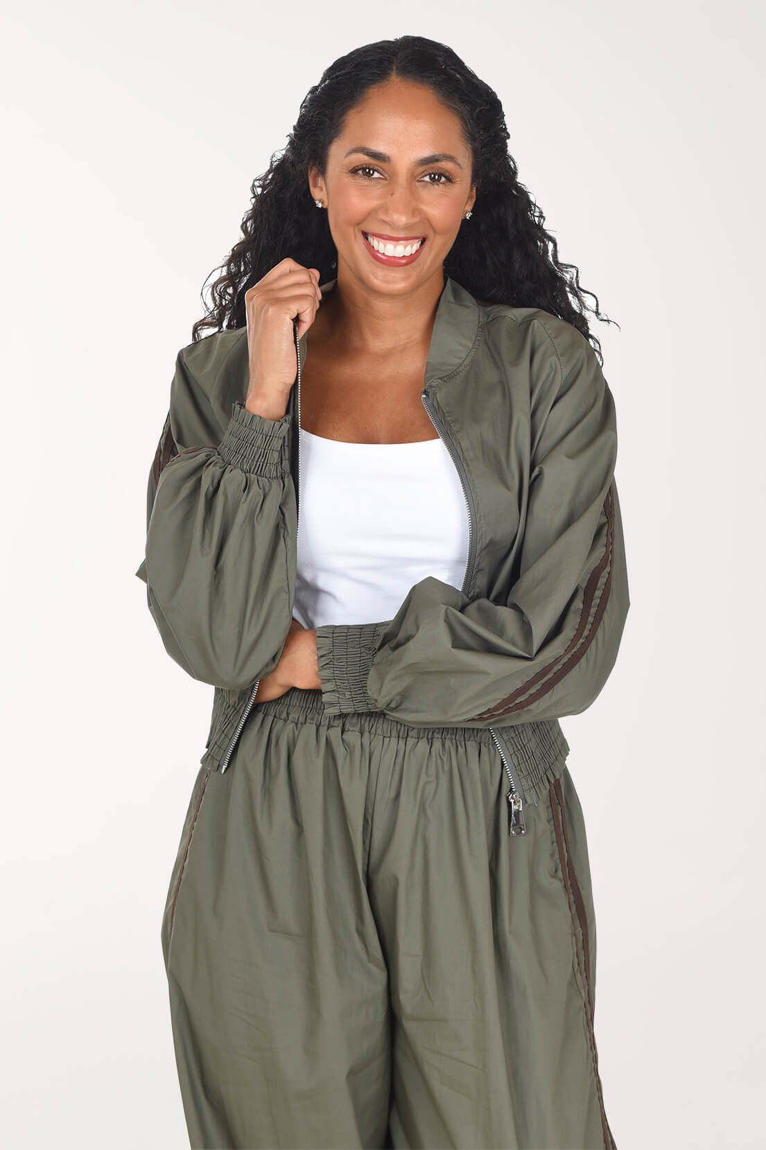 A smiling woman with long curly dark hair models an olive green bomber jacket and matching cargo pants over a white tank top, shot against a plain light background.