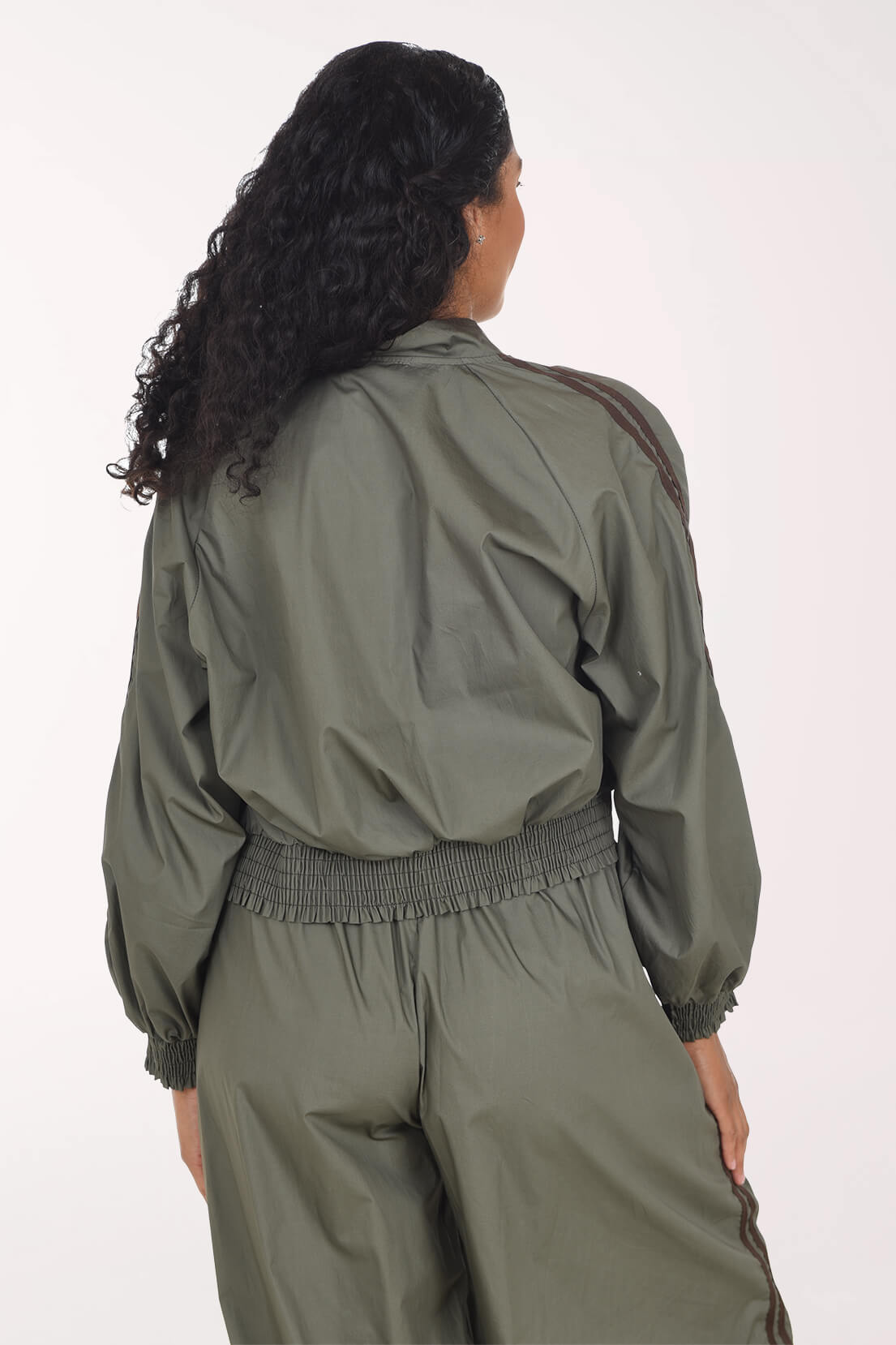 The back shot of a woman with long curly dark hair models an olive green bomber jacket and matching cargo pants shot against a plain light background.