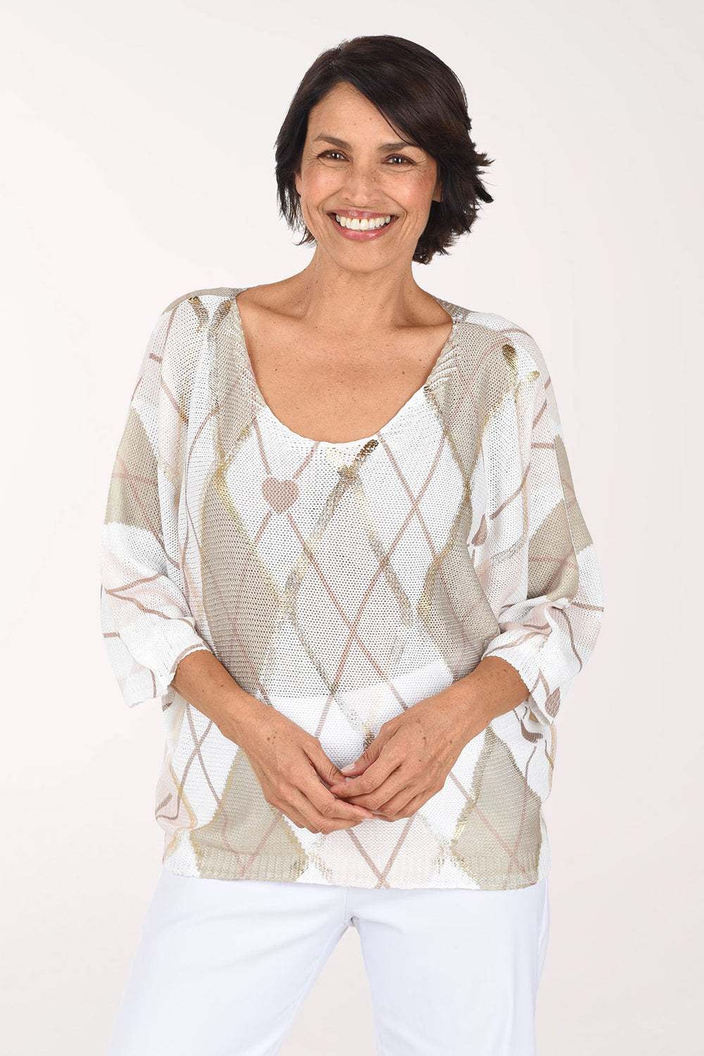 Smiling woman in an open-knit sweater with a wide crew neckline and three-quarter dolman sleeves, in a white and beige argyle print with small blush hearts and metallic gold accents, worn with white pants