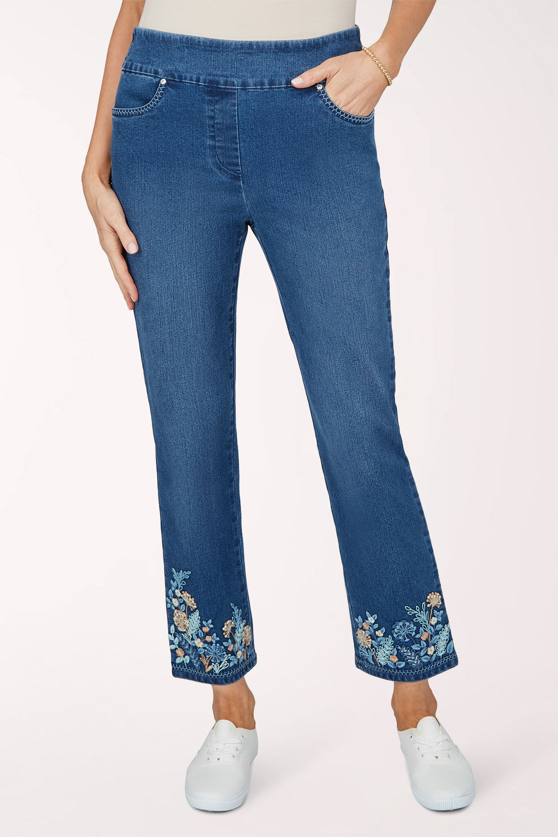 Front view of medium indigo stretch denim pull-on ankle pants with a slim leg and botanical embroidery in cream, peach and light blue around the hems, worn with white sneakers.