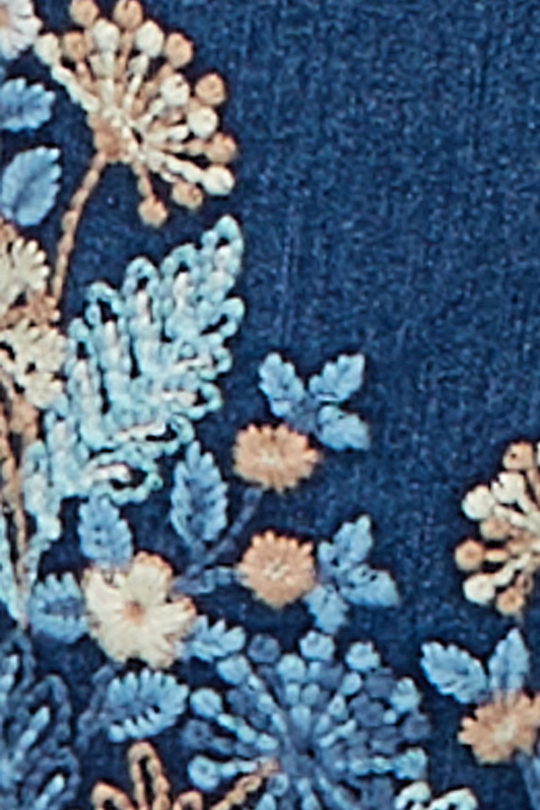 Close-up of the botanical embroidery on the hem of the denim pants, showing cream, peach and light blue flowers and leaves on medium indigo denim.