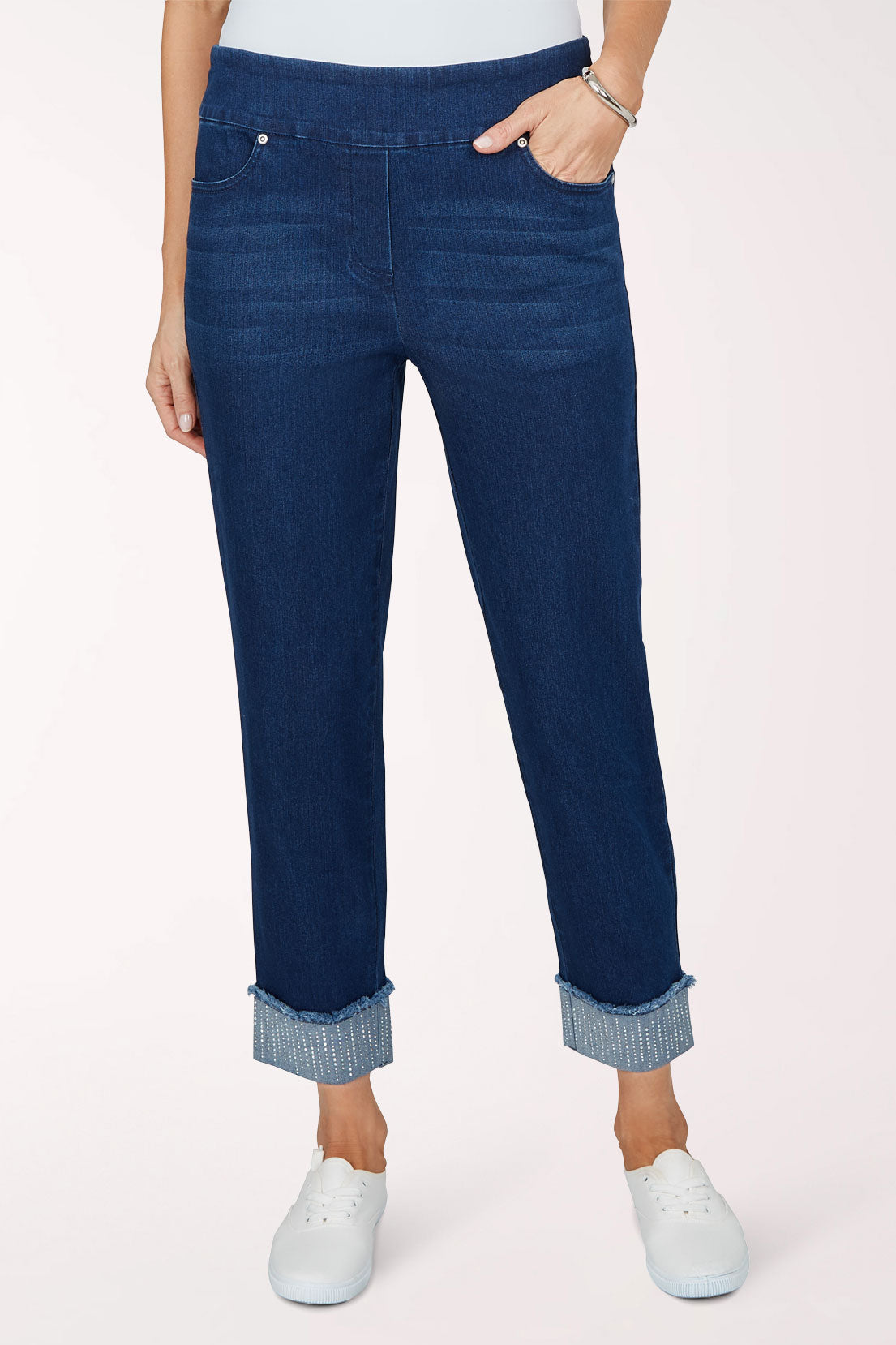 Front view of dark indigo stretch denim pull-on pants with a slim ankle-length leg and rolled cuffs trimmed with rhinestones, worn with white sneakers.