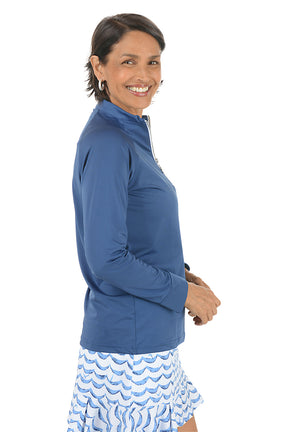 Woman wearing a blue long-sleeve top with a zipper, standing against a white background. Side shot.