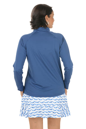 Woman wearing a blue long-sleeve top with a zipper, standing against a white background. Back shot.