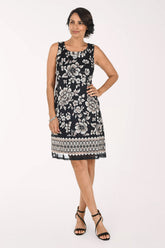 A woman modeling a sleeveless black and white floral print dress with a geometric patterned hem, paired with black strappy heels, against a plain white studio background.