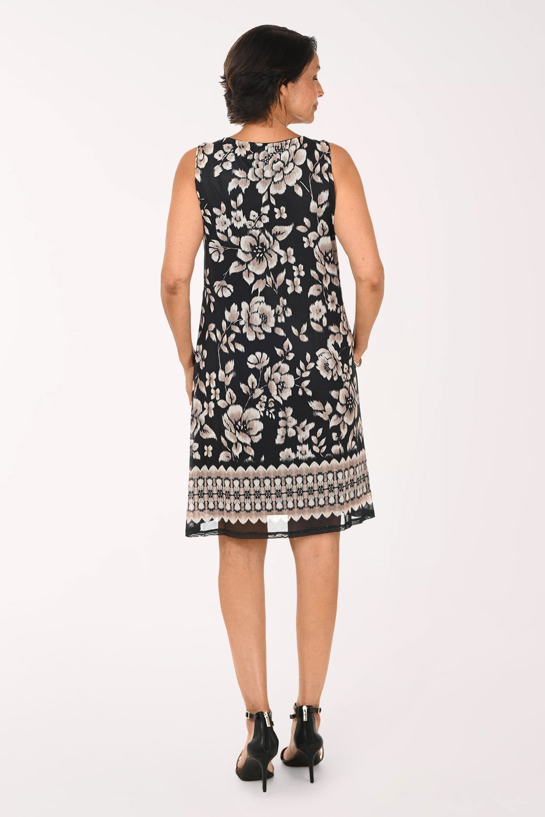 A woman modeling a sleeveless black and white floral print dress with a geometric patterned hem, paired with black strappy heels, against a plain white studio background.