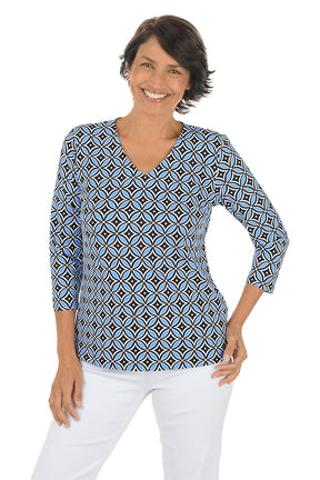 Woman wearing a geometric patterned blue and black blouse with white pants on a white background.