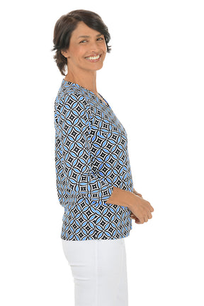 Woman wearing a geometric patterned blue and black blouse with white pants on a white background. Side shot.