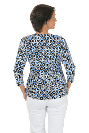Woman wearing a geometric patterned blue and black blouse with white pants on a white background. Back shot.