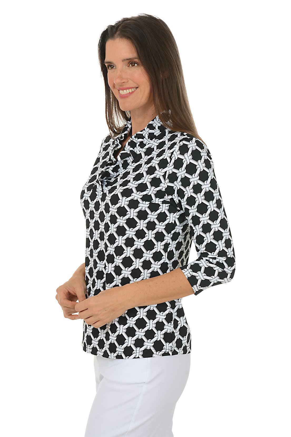 Woman wearing a black and white patterned blouse with white pants on a white background. Side shot.