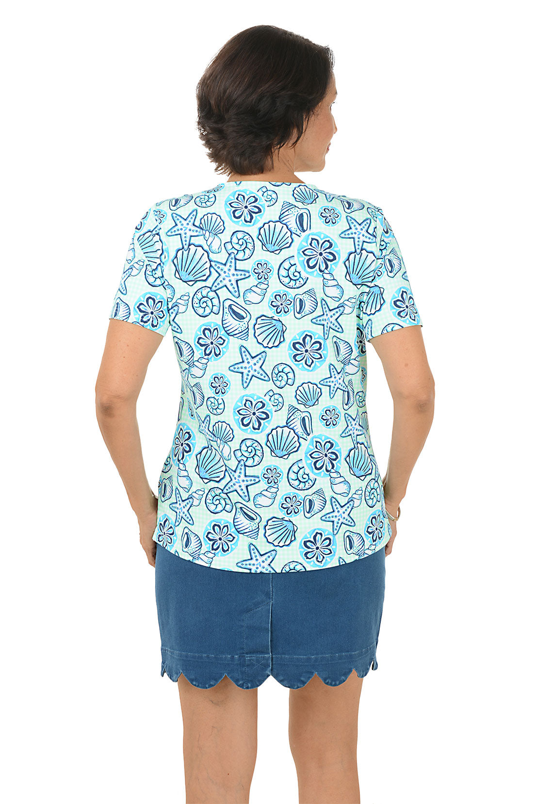 Woman wearing a blue patterned blouse with a shell design and a scallop denim skort on a white background. Back shot.