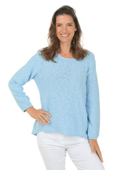 A woman wearing a long sleeve sky blue cotton scoop neck sweater, with a seam down the front and white pants.