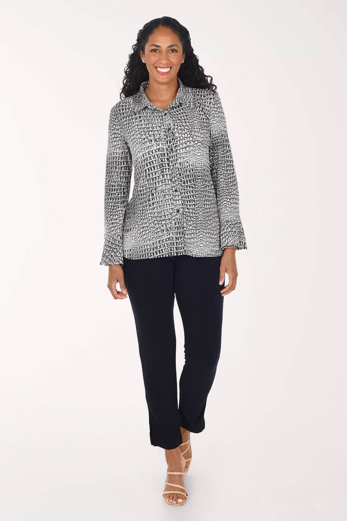 A woman modeling a long-sleeve button-up shirt with a black and white textured pattern, paired with navy pants and beige strappy heels, against a plain white studio background.