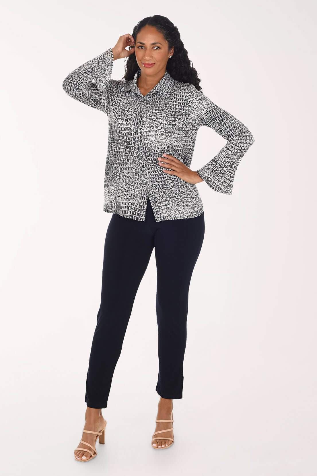 A woman modeling a black and white textured patterned long-sleeve button-up shirt with flared cuffs, paired with black pants and beige heeled sandals, against a plain white studio background.