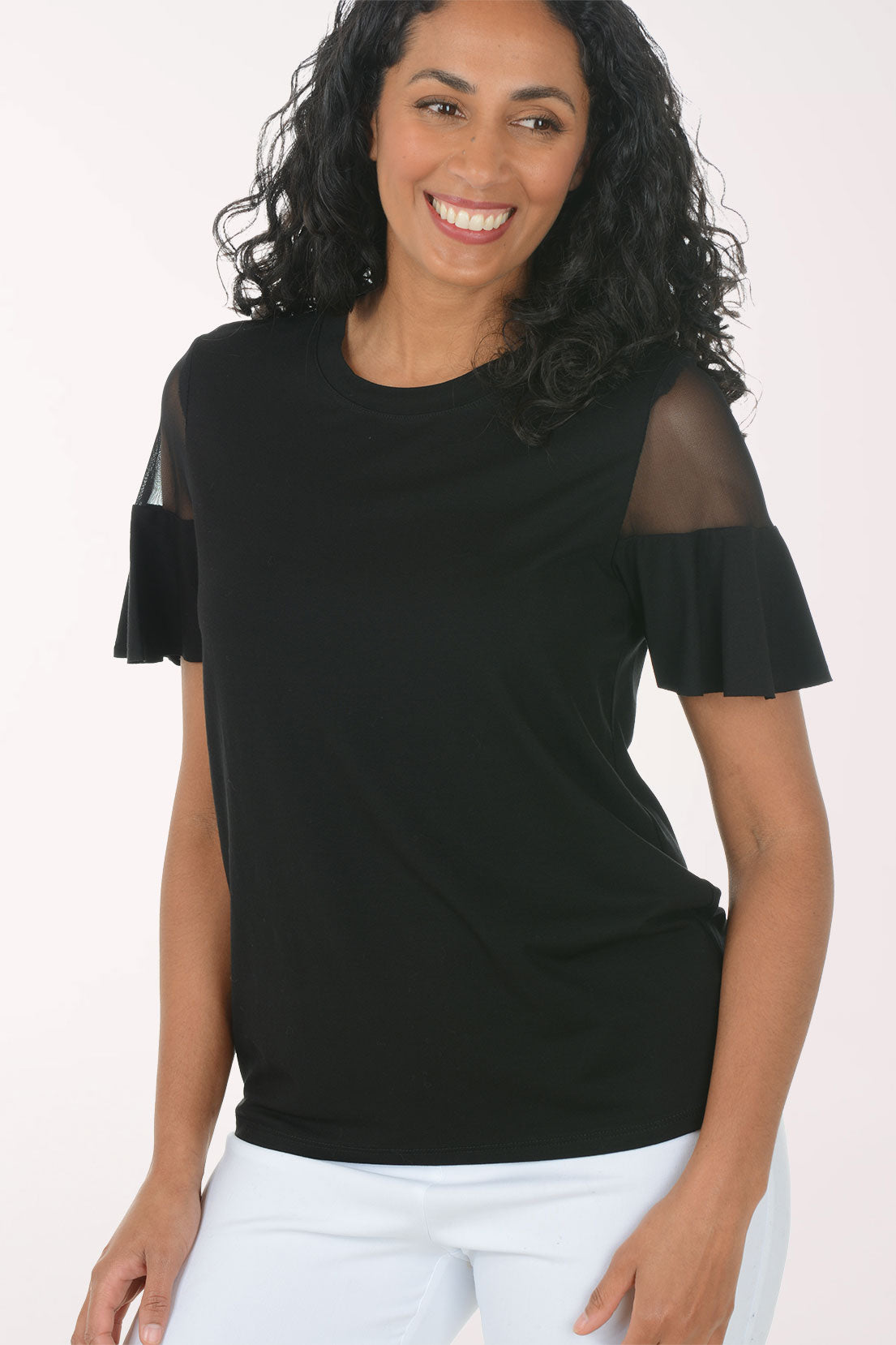 Woman wearing a black top with sheer sleeves and white pants on a light pink background