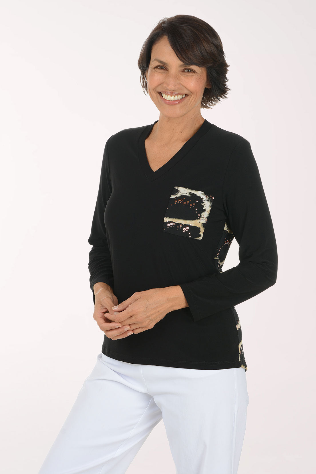 A smiling woman wearing a black long-sleeve V-neck sweater with a sequined abstract chest design in beige, white, and black, paired with white pants, against a plain white studio background.