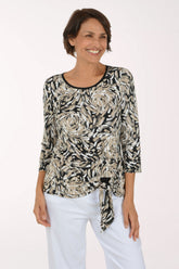 A woman modeling a three-quarter sleeve top with a black, white, and beige floral swirl pattern and a side tie, paired with white pants against a plain white background.