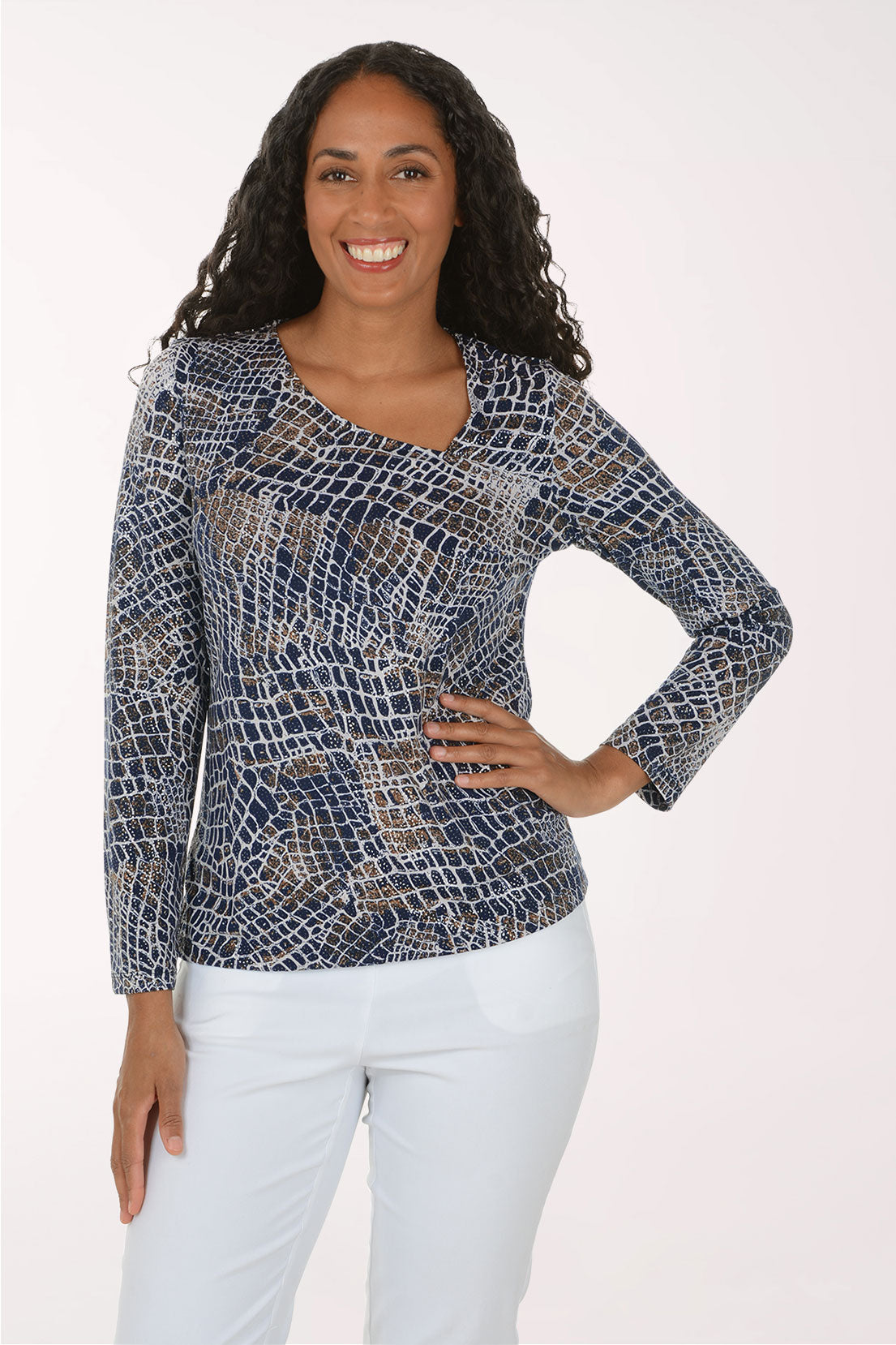 A woman modeling a long-sleeve V-neck top with a blue, white, and beige reptile or snakeskin pattern, paired with white pants, against a plain white studio background.